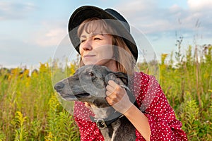 A young woman in a red dress hugs a greyhound dog outdoors and looks into the distance