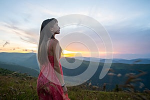 Young woman in red dress standing on grassy field on a windy evening in autumn mountains enjoying view of nature