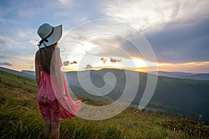 Young woman in red dress standing on grassy field on a windy evening in autumn mountains enjoying view of nature