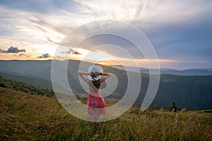 Young woman in red dress standing on grassy field on a windy evening in autumn mountains enjoying view of nature