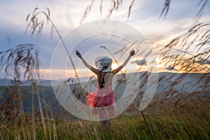 Young woman in red dress standing on grassy field on a windy evening in autumn mountains raising up her hands enjoying view of