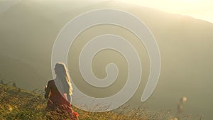 Young woman in red dress standing on grassy fild on a windy evening in autumn mountains enjoying view of nature.