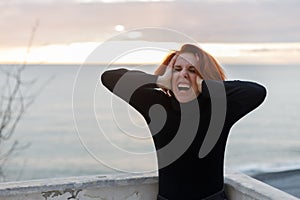 Young woman with red hair clutching her head in despair on the background of sea and sunset