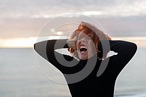 Young woman with red hair clutching her head and loudly screaming from heartache on the background of sea and sunset