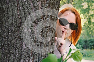 Young woman with red hair in sunglasses peeks out from behind a tree and shows gesture quietly