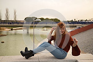 Young woman, red-haired, freckled, with red sweater and white headphones, sitting and dancing, listening to music, in an outdoor