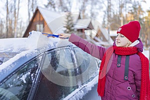 Young woman in a red hat cleaning a snow-covered car on the background of a winter road and the setting sun