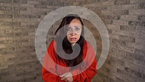 A young woman in a red hoodie is showing a fright on a brick background. A woman was gripped by fear.
