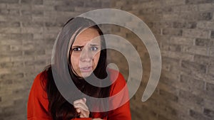 A young woman in a red hoodie is showing a fright on a brick background. A woman was gripped by fear.