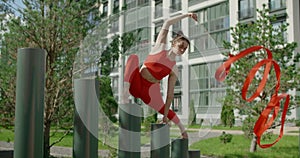 Young woman in red performs callisthenics with ribbon in the libing apartment yard, gymnast does acrobatic exercises in