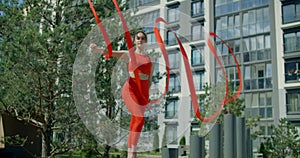 Young woman in red performs callisthenics with ribbon in the libing apartment yard, gymnast does acrobatic exercises in