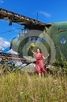 Young woman in a red pin up dress waving a bunch of flowers to someone at an abandoned airplane