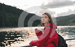 Young woman in a red raincoat looking at the camera, smiling, enjoying staying in the woods near the lake. Cute attractive girl