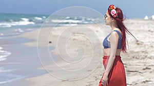 Young woman in a red skirt and blue top with red hair flying in the wind and a floral wreath in her hair against the