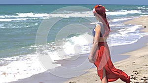 Young woman in a red skirt and blue top with red hair flying in the wind looks at the stormy sea