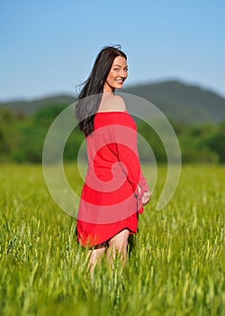 Young woman in red summer dress walking in green unripe wheat field, looking back over her shoulder