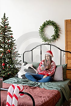 Young woman in red sweater and santa hat sitting on the bed using mobile phone texting or having video call