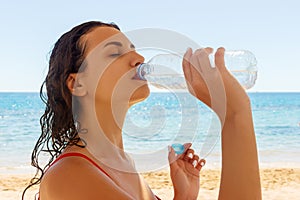 Young woman in a red swimsuit, drinking sparkling water from a transparent bottle on the beach