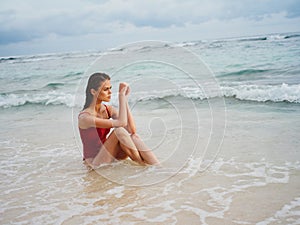 A young woman in a red swimsuit sits on the sand with a beautiful sun tan and looks out at the ocean in the tropics on