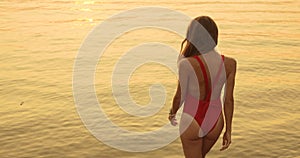 Young woman in red swimsuit is walking on sea at golden sunset, back view.