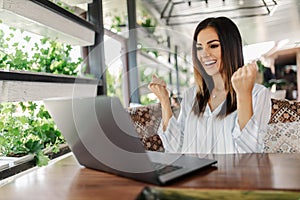 Young woman rejoices in winning having raised her hands up sitting in front of laptop in cafe