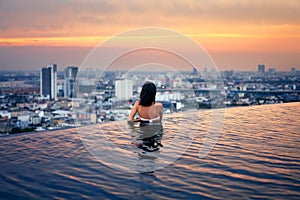 Young woman relax in swimming pool on roof top during amazing sunset and enjoy city view