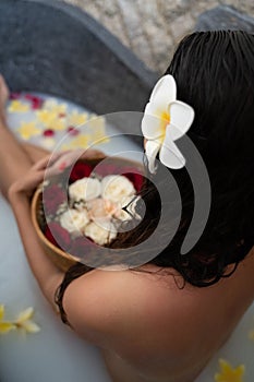 Young woman relaxing in black stone bath with tropical flowers and rose petals. Skin treatment, luxury spa concept