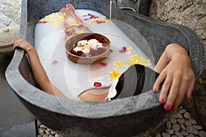 Young woman relaxing in black stone bath with tropical flowers and rose petals. Skin treatment, luxury spa concept
