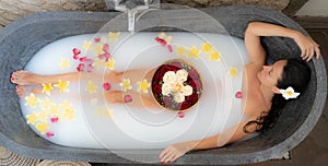 Young woman relaxing in black stone bath with tropical flowers and rose petals. Skin treatment, luxury spa concept