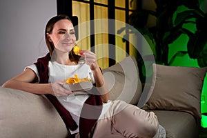 Young woman is relaxing on the couch, watching TV and eating chips