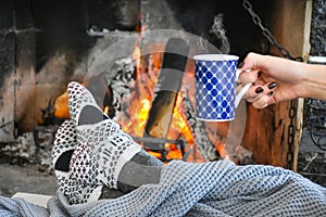 Young woman relaxing in front of cozy fireplace and warming up her feet in woolen socks in country house.