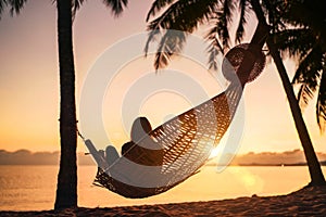 Young woman relaxing in hammock hinged between palm trees on the sand beach at orange sunrise morning time