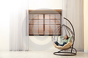 Young woman relaxing in hanging chair near window. Space for text