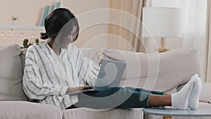 Young woman relaxing at home on cozy sofa with laptop chatting with friends in social network smiling typing email makes