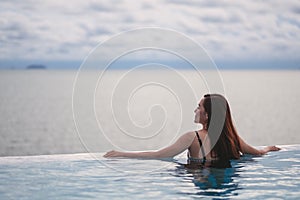 A young woman relaxing in infinity swimming pool looking at a beautiful sea view