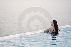 A young woman relaxing in infinity swimming pool looking at a beautiful sea view