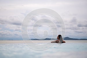 A young woman relaxing in infinity swimming pool looking at a beautiful sea view