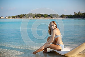 Young woman relaxing in a modern deck chair on a tropical beach with glasses on. Girl is sitting on a beach sun bed