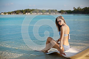 Young woman relaxing in a modern deck chair on a tropical beach with glasses on. Girl is sitting on a beach sun bed