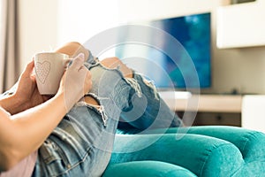 Young woman relaxing on the sofa at home, watching tv and enjoying coffee