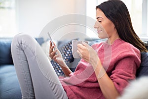 Young Woman Relaxing On Sofa With Hot Drink Checking Mobile Phone