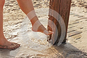 Young woman removing sand form her legs under beach shower near sandy ground, water drops spraying over feet