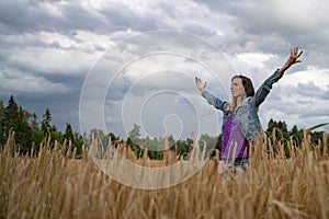 Young woman with a reserved worried expression on her face standing in the middle of golden wheat field with her arms spread wide