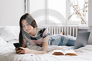 Young woman resting on a bed at home and using laptop computer and smartphone