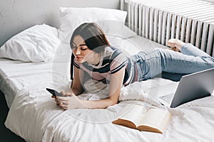 Young woman resting on a bed at home and using laptop computer and smartphone