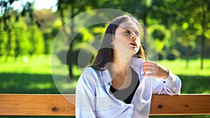 Young woman resting on bench in park suffering from heat and stuffiness, pms
