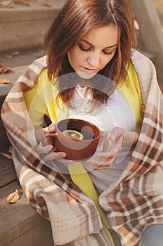 Young woman resting and drinking tea sitting in autumn garden on the steps, wrapped in a woolen plaid blanket.