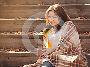 Young woman resting and drinking tea sitting in autumn garden on the steps, wrapped in a woolen plaid blanket.