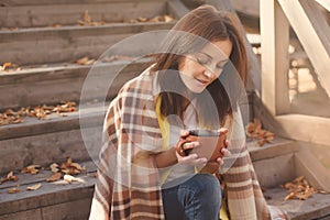 Young woman resting and drinking tea sitting in autumn garden on the steps, wrapped in a woolen plaid blanket.