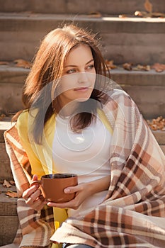 Young woman resting and drinking tea sitting in autumn garden on the steps, wrapped in a woolen plaid blanket.